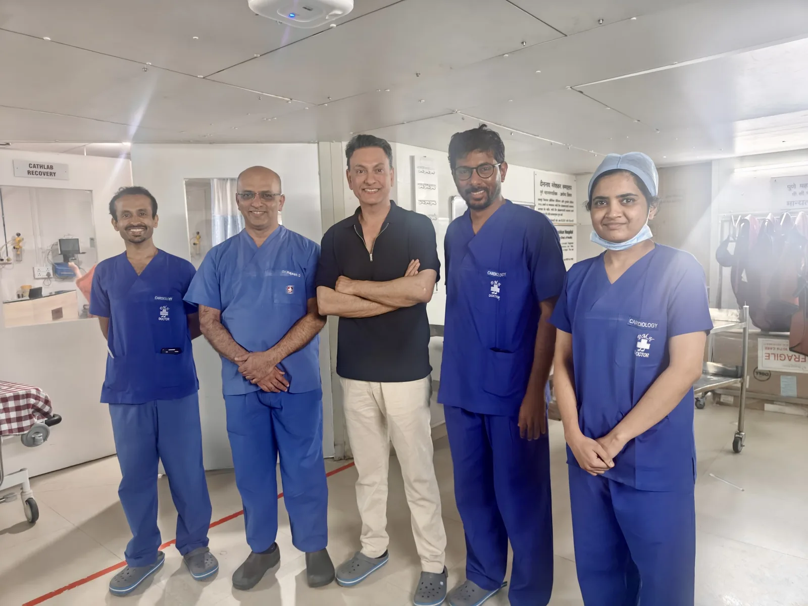 Five doctors stand together in a bright cath lab recovery bay, name badges reading cardiology, with Professor Gupta in the centre out of scrubs.