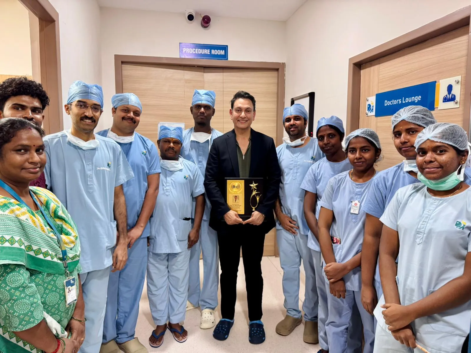 A dozen staff in pale blue scrubs and theatre caps, and one in a green sari, gather round Professor Gupta, in a dark suit, as he holds a gilt plaque outside a door marked Procedure Room.