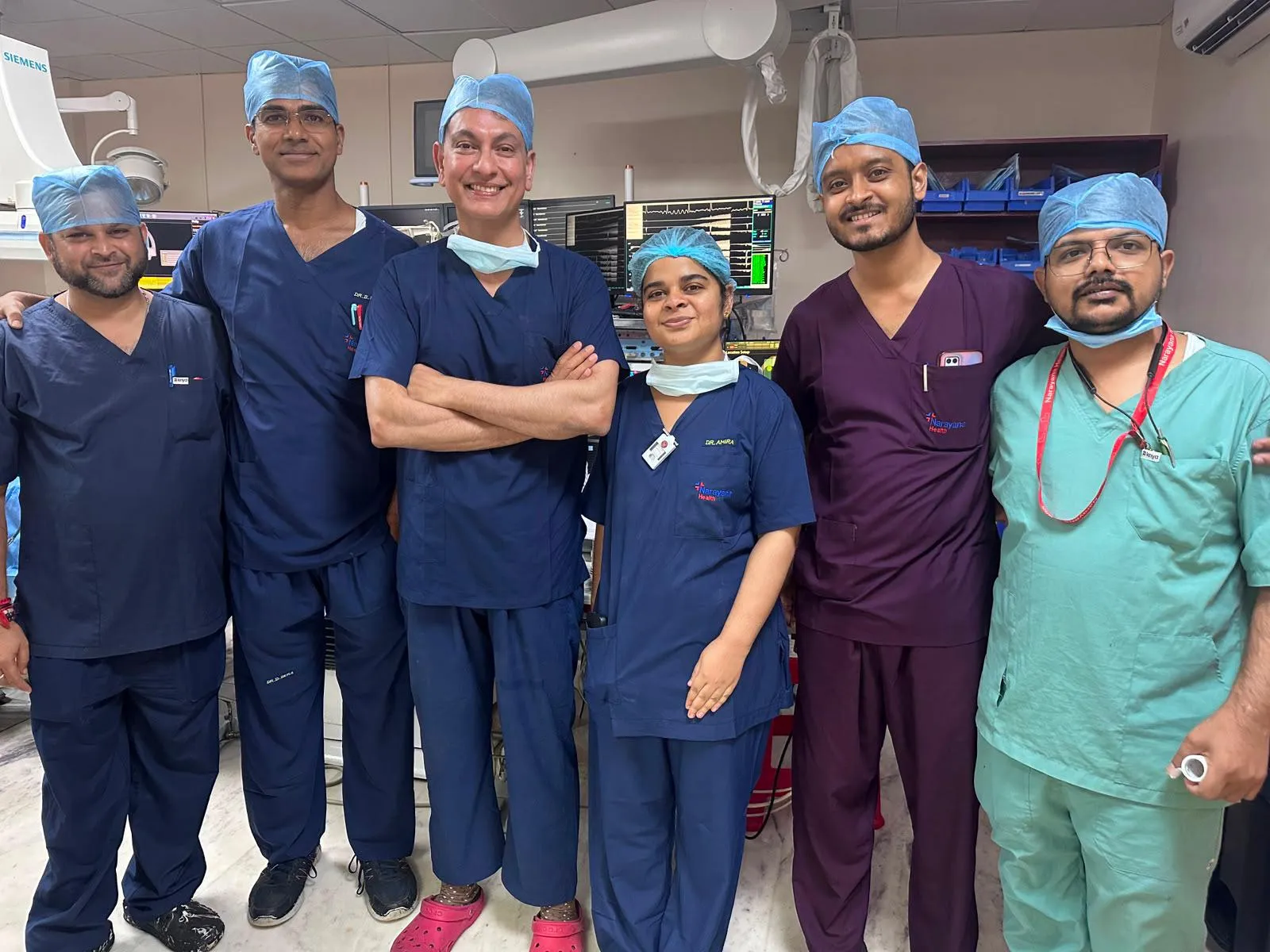 Six clinicians in navy, maroon and green scrubs and theatre caps stand arm in arm in front of a bank of cath lab monitors showing live electrograms, Professor Gupta in the centre with his arms folded.