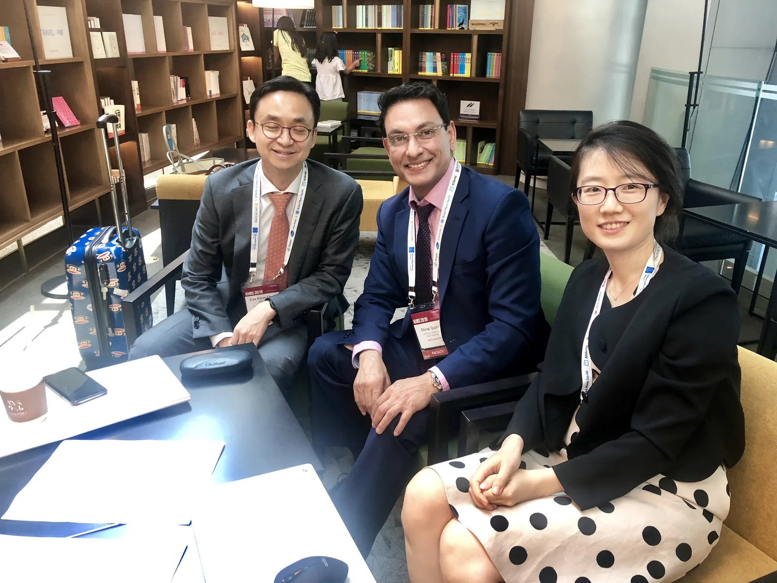 Professor Gupta sits between two Korean colleagues in a conference lounge, all three wearing delegate badges, bookshelves and meeting seating behind them.