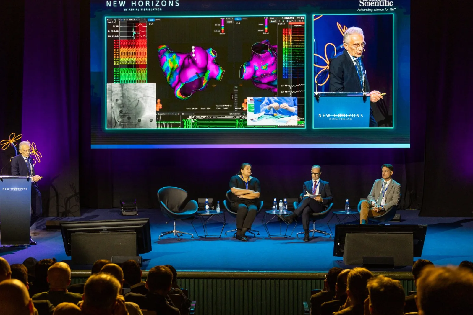 A lit stage in front of a full auditorium. Three faculty sit in armchairs while a fourth speaks at a lectern, under a wide screen showing a colour 3D voltage map of the left atrium alongside live electrograms.