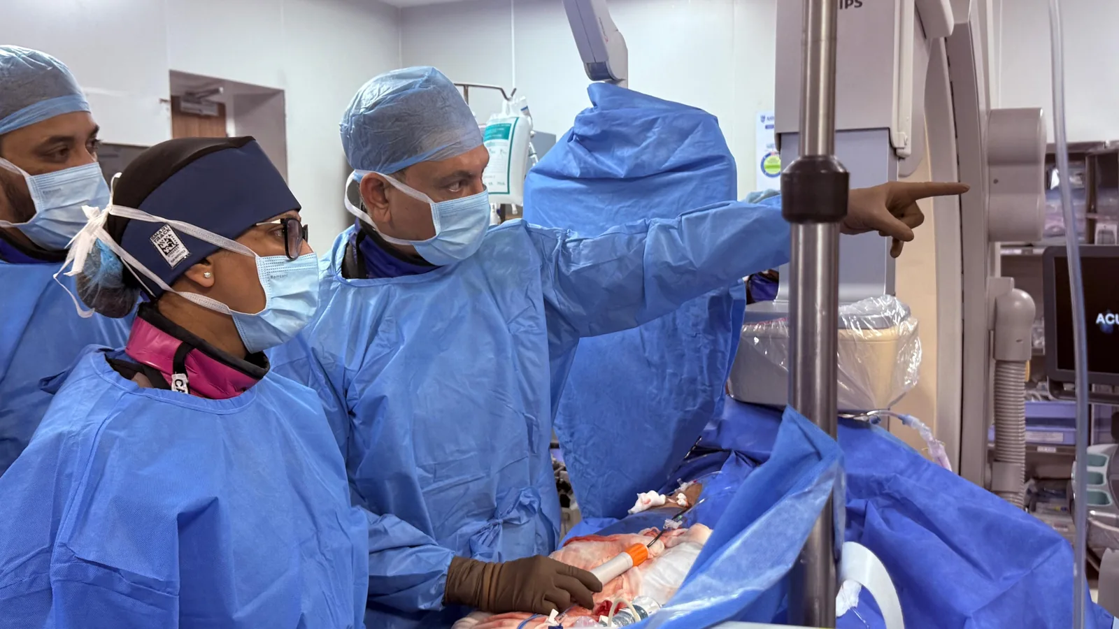 Professor Gupta, gowned and masked at the ablation table, points to the mapping screen while a colleague beside him follows the direction. The X-ray C-arm and the draped patient are in view.